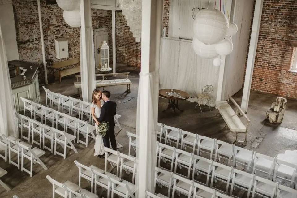 Image of a couple embracing in the center of a rustic-chic interior that's been beautifully set up for a wedding ceremony