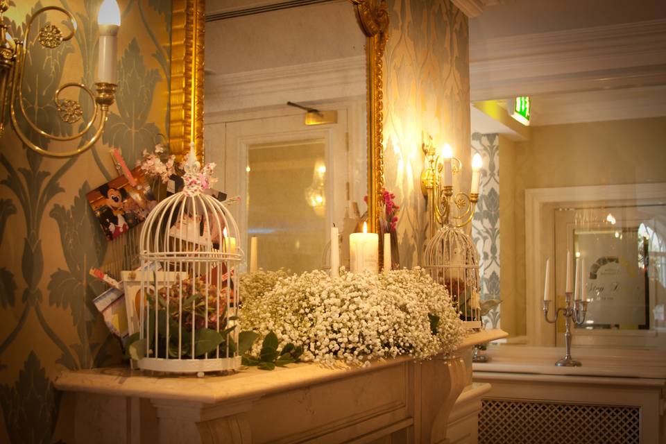 A vintage white birdcage sits on a vanity surrounded by flowers, wall sconces, and a large gold leaf mirror