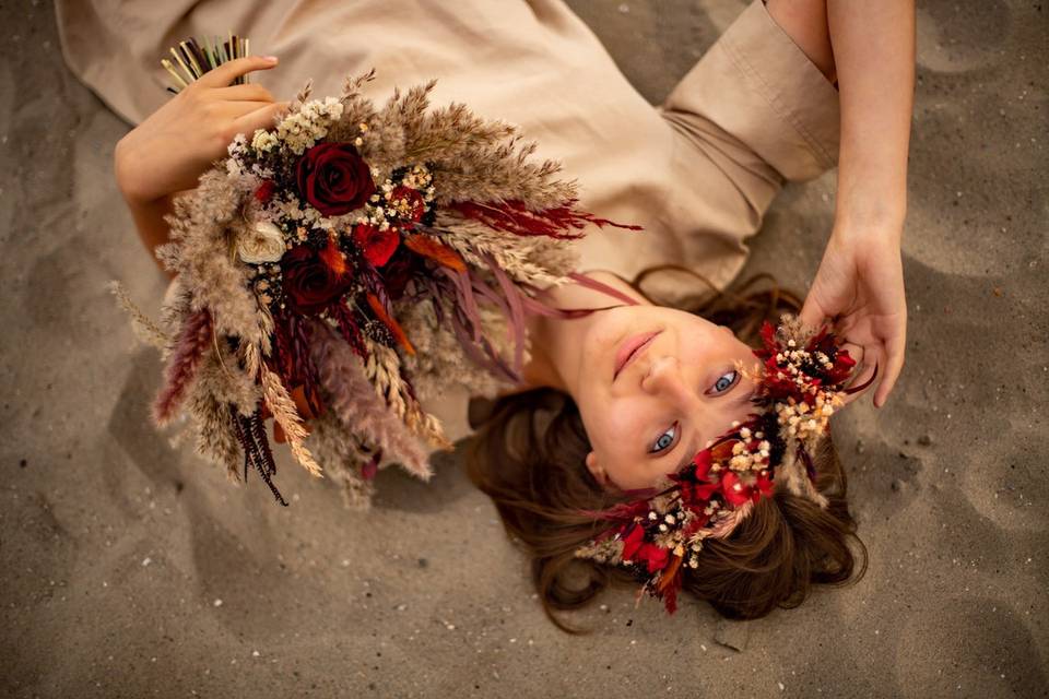 Woman lying in sand wearing a flower crown made with red flowers and other foliage, holding a coordinating bouquet of dried flowers