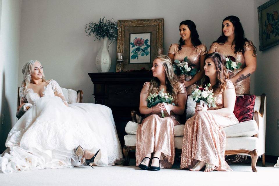 On the left side of the photo, a bride in a wedding dress sits back in a chair. The bride is looking at four bridesmaids in matching sequin pink dresses.