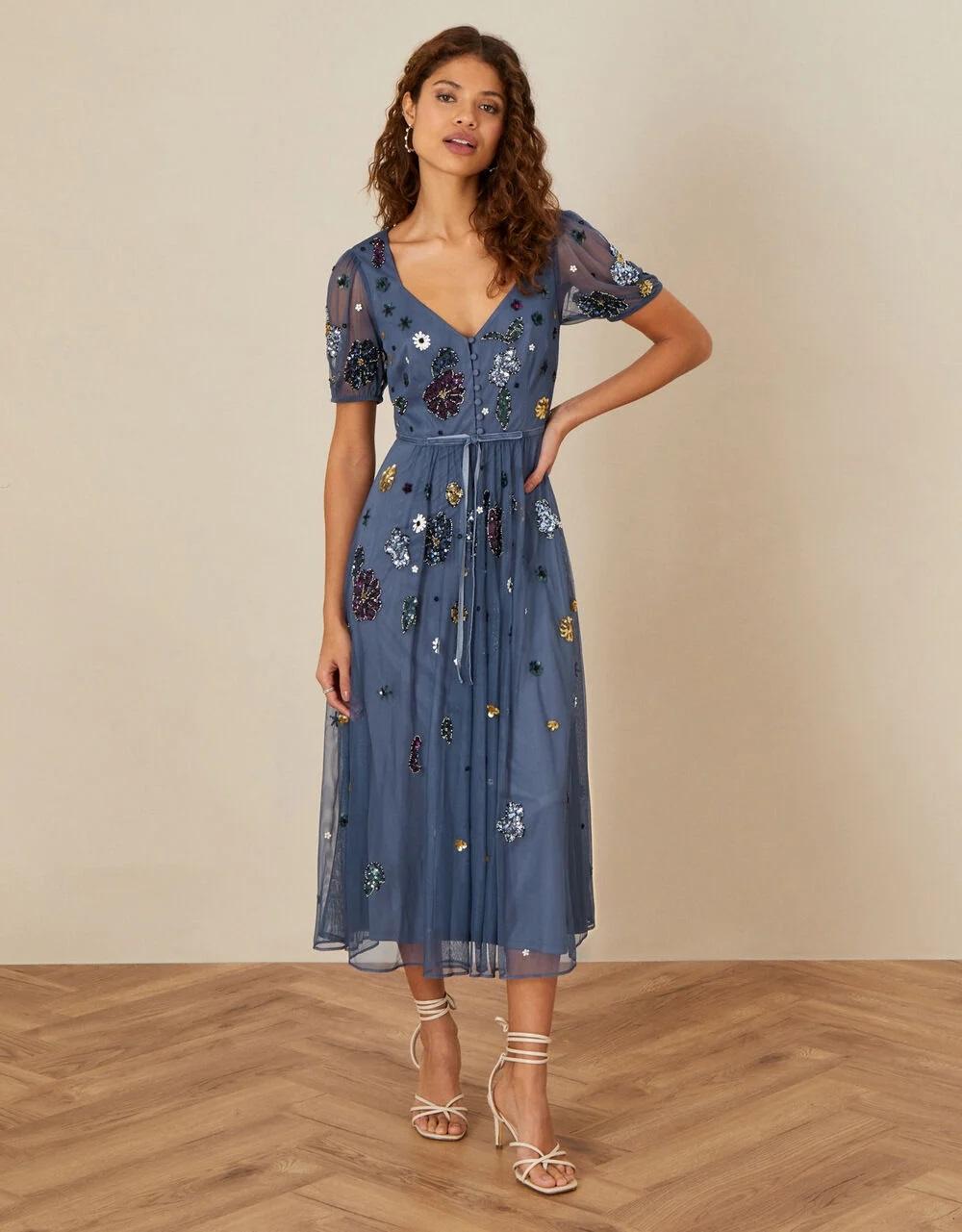 Model is standing hand-on-hip wearing a blue dress with flowers on it.