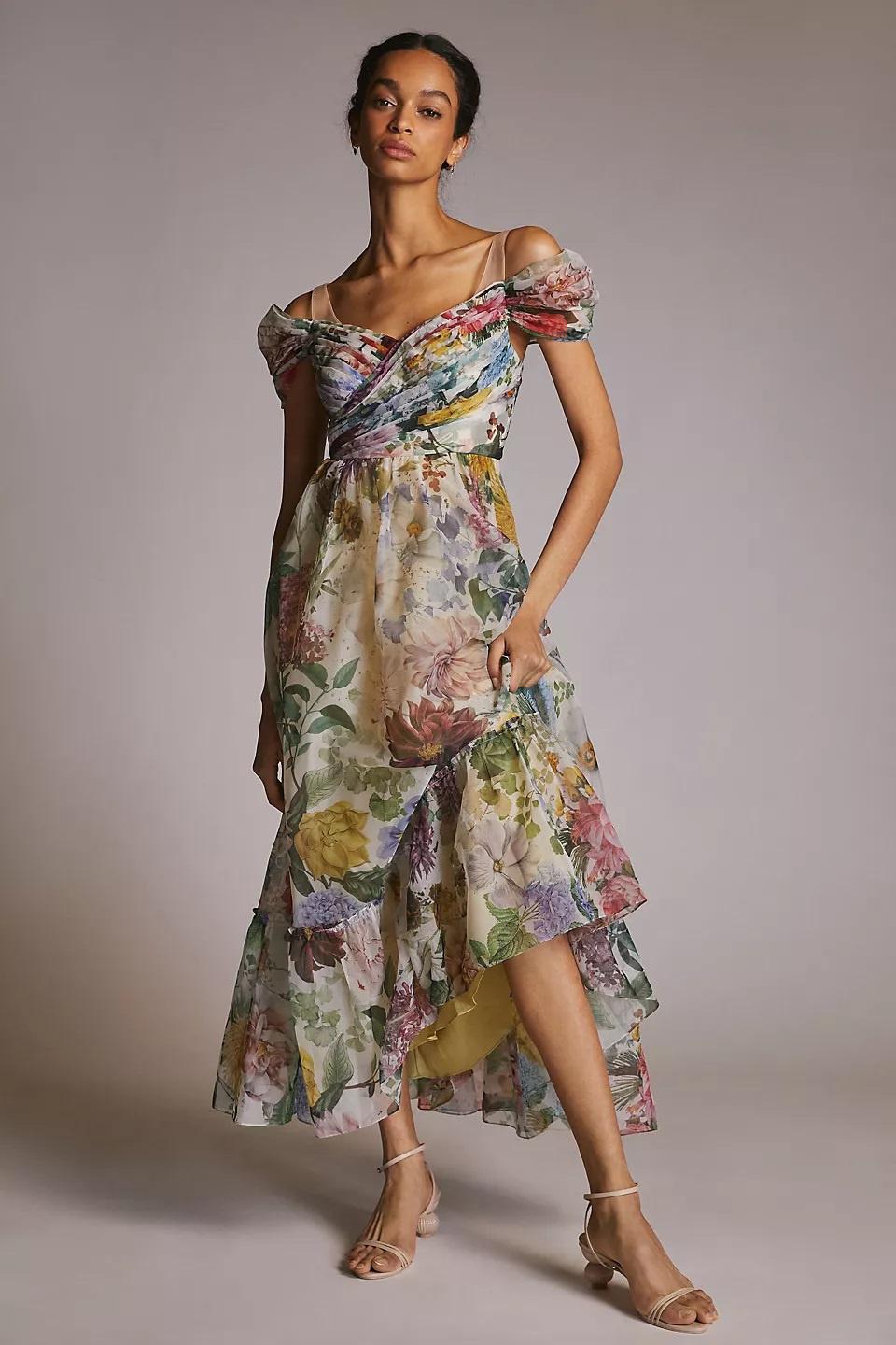 Model wearing a dress with floral design throughout. The puffed sleeves are off-the-shoulder and the skirt has a ruffle towards the hem.